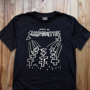 Może przedstawiać: Czarny T-shirt z białym tekstem, który brzmi "Maitre des Marionnettes" i grafiką trzech krzyży z promieniami światła nad nimi. Tekst "Les Arcanes Qui Dorment" jest nadrukowany mniejszą czcionką nad głównym tekstem.
