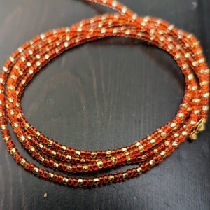 May include: A coiled orange beaded waist bead strand with gold-colored accents. The beads are small and evenly spaced, creating a textured appearance. The strand is designed to be worn around the waist.