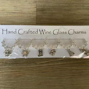 May include: Set of six silver wine glass charms with Christmas-themed designs. The charms feature a gift box, a star, a stocking, a bell, a Christmas tree, and a snowman. The charms are attached to a string of white beads.
