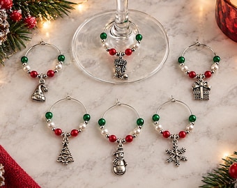 May include: A set of six silver-toned metal wine glass charms featuring holiday-themed pendants including a stocking, gift box, Christmas tree, snowman, snowflake, and hat. Each circular charm is accented with red, white, and green beads.