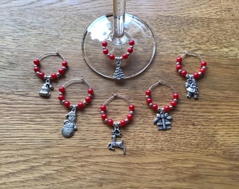 May include: Six red and silver Christmas themed wine glass charms. Each charm features a different design, including a snowman, a reindeer, a Christmas tree, a gift box, a Santa Claus, and a stocking.