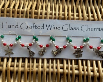 May include: Set of six hand-crafted wine glass charms featuring Christmas-themed designs. Each charm is made with red, white, and green beads and a silver charm. The charms include a snowflake, a Christmas tree, a Santa hat, a stocking, a gift box, and a snowman.