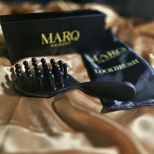 May include: Black hairbrush with rounded bristles, a handle, and the text "LOCKBRUSH" on a black satin bag. The brush and bag are on a gold satin surface. The box in the background has the text "MARQ".