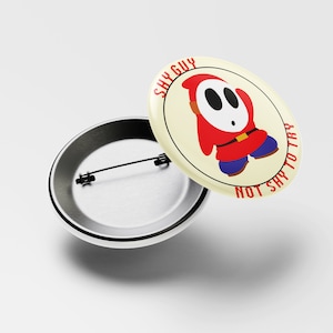 May include: A round button with a white background and a red and blue Shy Guy character from the Super Mario Bros. video game series. The text "Shy Guy Not Shy To Try" is written around the character.