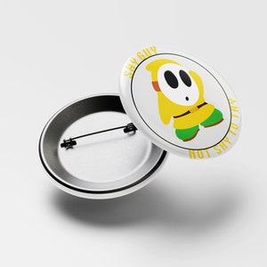 May include: A white button with a yellow Shy Guy character from the Super Mario video game series. The text on the button reads "Shy Guy Not Shy To This".