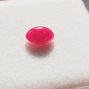 May include: A polished, oval ruby gemstone in a vivid red hue. The gem is displayed on a white, textured surface. The stone's smooth surface reflects light, creating a bright appearance.