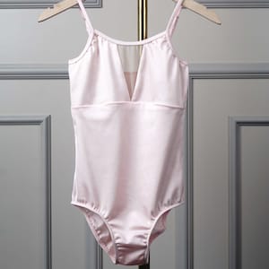 Könnte beinhalten: Hellrosa Satin-Leotard mit V-Ausschnitt und Spaghettiträgern. Das Leotard hat einen transparenten Mesh-Einsatz am Ausschnitt und hängt an einem Holzbügel. Geeignet für Tanz oder Gymnastik.