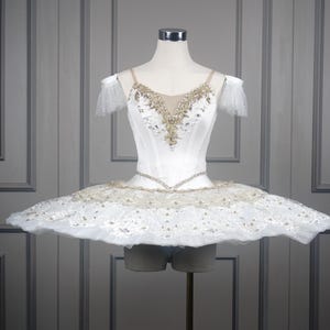 Może przedstawiać: Biała baletowa tutu z gorsetem ozdobionym złotymi zdobieniami i przezroczystym dekoltem. Tutu ma pełną, warstwową spódnicę z delikatną koronką i złotymi akcentami. Kostium jest prezentowany na manekinie.