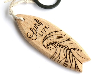 May include: Wooden surfboard keychain with a wave design and the text "Surf Life". The keychain has a black metal ring and a white string.