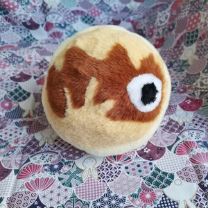 Könnte beinhalten: Ein Plüschtier in Form einer Kugel mit einem braunen und weißen Fischdesign. Das Spielzeug ist aus weichem, flauschigem Stoff und hat eine hellbraune Farbe.