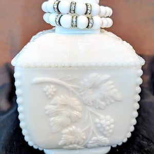 May include: A white, square-shaped trinket box with a lid, featuring a raised grape and leaf design. The box is adorned with a beaded edge and topped with two white beaded bracelets with rhinestone accents. The overall aesthetic is vintage and decorative.