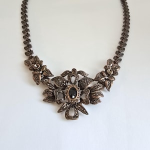 May include: A necklace featuring a dark metallic chain with a repeating leaf-like pattern. The centerpiece is an ornate floral design encrusted with small dark stones and a single oval black stone at the center, flanked by two smaller floral accents.