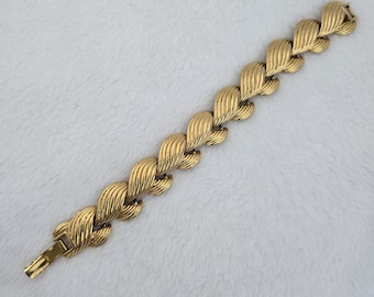 May include: A gold-tone metal bracelet featuring a series of interconnected, textured, wave-like links. The design consists of repeating, curved segments with fine, parallel ridges, finished with a secure, rectangular clasp at one end.