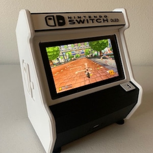 Könnte beinhalten: Eine weiße und schwarze Nintendo Switch OLED-Konsole in einem benutzerdefinierten Arcade-Schrank. Der Schrank hat eine schwarze Oberseite mit dem Nintendo Switch OLED-Logo und dem Text "NINTENDO SWITCH OLED". Der Schrank ist weiß mit einem schwarzen Sockel.