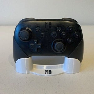 以下が含まれることがあります： 白い3Dプリントされたスタンドに置かれた黒いニンテンドースイッチプロコントローラー。スタンドの前面にはニンテンドースイッチのロゴがあります。
