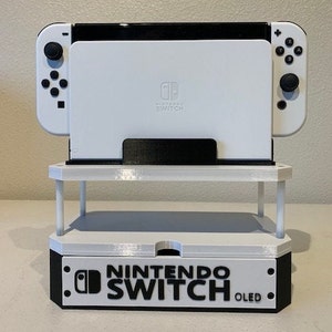 May include: White Nintendo Switch OLED console with a white stand. The console is on a white base with the words "NINTENDO SWITCH OLED" printed in black.