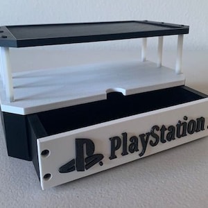 Peut inclure: Un organisateur à thème PlayStation noir et blanc avec un tiroir et une étagère supérieure. Le tiroir a un devant blanc avec le logo PlayStation et le mot "PlayStation" imprimé en noir.