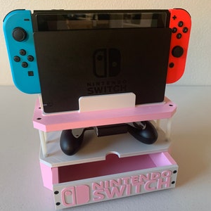 Pode incluir: Um suporte rosa para Nintendo Switch com uma gaveta. O suporte tem uma prateleira superior branca para o console e uma prateleira inferior para os controladores. A gaveta tem uma frente branca com o logotipo e o texto Nintendo Switch.