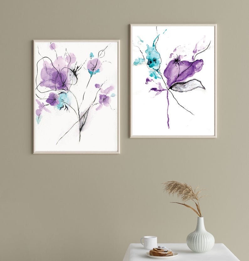 May include: Two framed watercolor floral art prints. The artwork features abstract purple and teal flowers with black line details on a white background. The frames are a light neutral color.