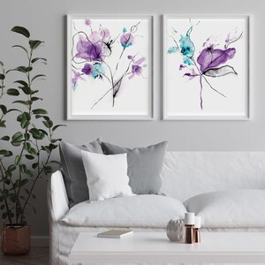 May include: Two framed floral art prints. Each print features abstract watercolor flowers in shades of purple and teal, with black line details. The artwork is displayed on a white background within a white frame. The prints are displayed above a white sofa.