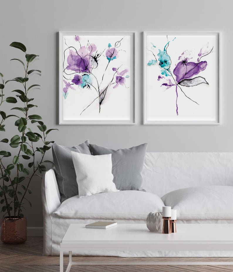 May include: Two framed floral art prints. Each print features abstract watercolor flowers in shades of purple and teal, with black line details. The artwork is displayed on a white background within a white frame. The prints are displayed above a white sofa.