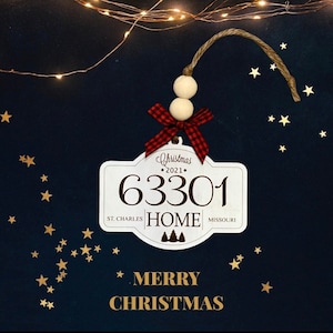 Puede incluir: Adorno navideño de madera blanco con el texto "Christmas 2021" y el número "63301" con las palabras "ST. CHARLES HOME MISSOURI" debajo. El adorno tiene un lazo de cuadros rojos y negros y cuentas de madera colgando de la parte superior. El texto "MERRY CHRISTMAS" está escrito en dorado debajo del adorno.
