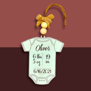 May include: A light blue baby onesie ornament with the name Oliver, birth weight of 6 lbs 5 oz, birth length of 19 inches, and birth date 6/16/2021. The ornament has a brown twine hanger with a brown bow and two wooden beads.