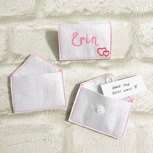 May include: Two white felt envelopes with pink stitching. One envelope contains a white card with the text "Have the best day!" and a pink heart. The other envelope has a white felt rectangle with the name "Erin" embroidered in pink with two pink hearts.