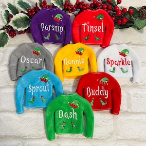 May include: Seven elf-sized Christmas jumpers in various colours, including purple, red, grey, yellow, white, blue, and green. Each jumper has a different name embroidered on it, including Parsnip, Tinsel, Oscar, Ronnie, Sparkle, Sprout, Buddy, and Dash.