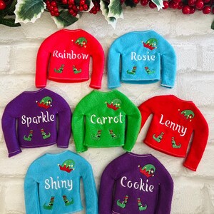 May include: Six elf-sized Christmas sweaters in different colors with personalized names: Rainbow, Rosie, Sparkle, Carrot, Lenny, and Cookie. Each sweater features a small elf hat and shoes.