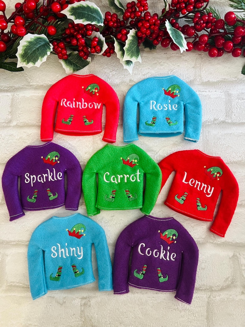 May include: Six elf-sized Christmas sweaters in different colors with personalized names: Rainbow, Rosie, Sparkle, Carrot, Lenny, and Cookie. Each sweater features a small elf hat and shoes.