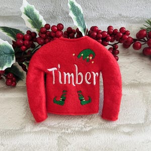 May include: Red felt elf sweater with green elf hat and shoes embroidered on the front. The sweater has the name "Timber" embroidered on the front.