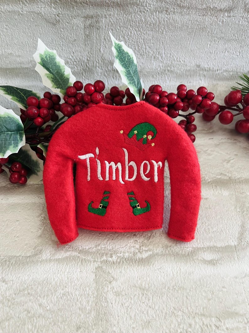 May include: Red felt elf sweater with green elf hat and shoes embroidered on the front. The sweater has the name "Timber" embroidered on the front.
