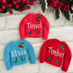 May include: Three elf-themed Christmas sweaters with personalized names: Tinsel, Elvis, and Timber. The sweaters are red and blue with white lettering and elf hat and shoe graphics.