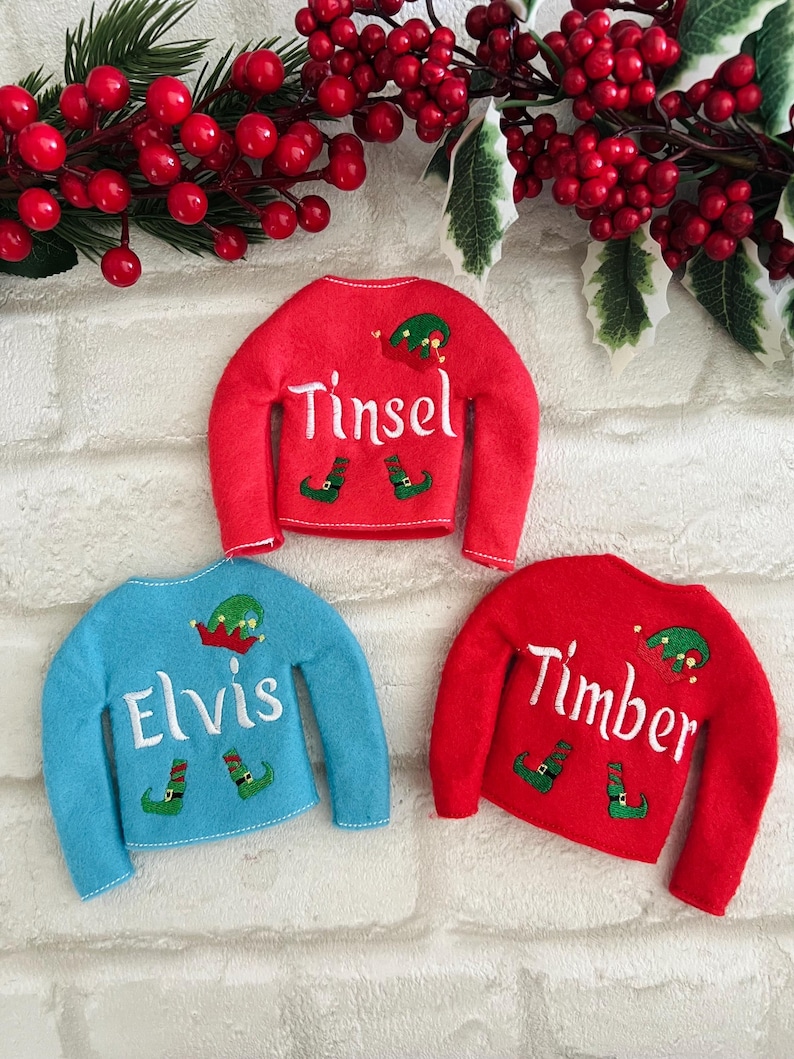 May include: Three elf-themed Christmas sweaters with personalized names: Tinsel, Elvis, and Timber. The sweaters are red and blue with white lettering and elf hat and shoe graphics.