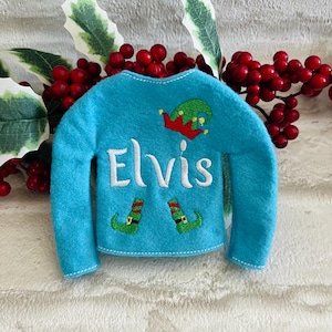 May include: A blue elf sweater with the name "Elvis" embroidered on the front. The sweater has a green elf hat and two green elf shoes embroidered on the front.