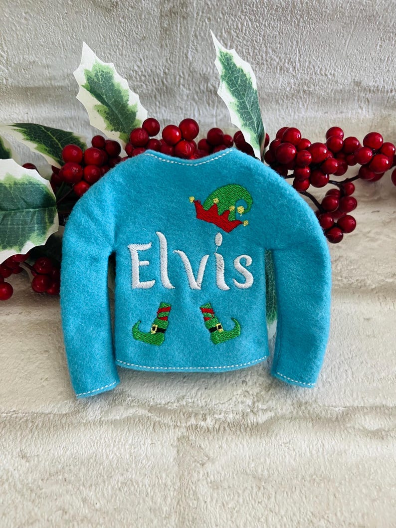 May include: A blue elf sweater with the name "Elvis" embroidered on the front. The sweater has a green elf hat and two green elf shoes embroidered on the front.