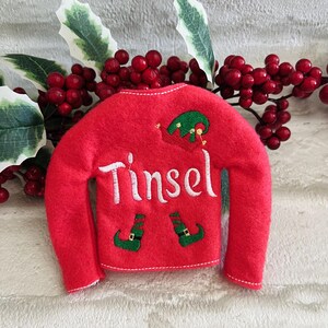May include: Red felt elf stocking with white embroidery that reads "Tinsel" and green elf shoes.