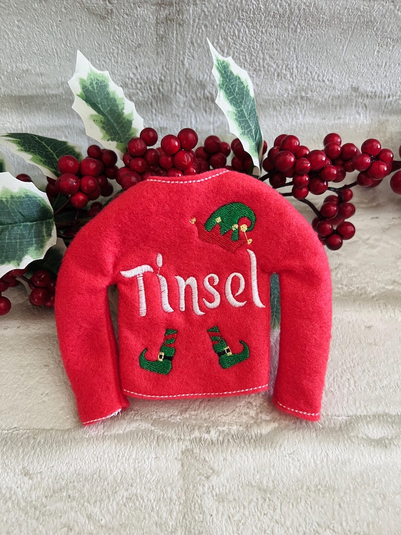 May include: Red felt elf stocking with white embroidery that reads "Tinsel" and green elf shoes.