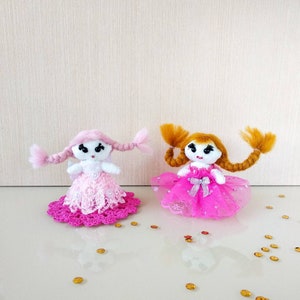 Op de afbeelding: Twee handgemaakte poppen met witte gezichten, elk met haar van garen in vlechten. De ene pop draagt een roze kanten jurk, de andere een roze tule jurk. Beide poppen hebben een strik en staan op een reflecterend oppervlak.
