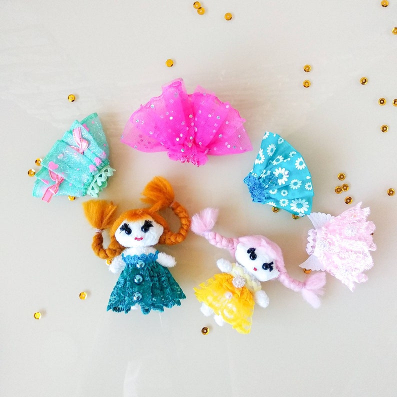 Op de afbeelding: Handgemaakte poppenfiguren met garen haar en stoffen jurken in teal, geel en roze. De poppen hebben geschilderde gezichten en zijn omringd door kleine gouden confetti stukjes. De jurken zijn versierd met kant en pailletten.