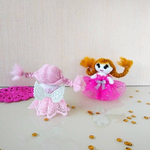 Op de afbeelding: Twee handgemaakte poppen. E&eacute;n pop is roze met witte vleugels en een kanten jurk. De andere pop heeft oranje haar en een roze jurk met een zilveren strik. Gouden confetti is verspreid rond de poppen.