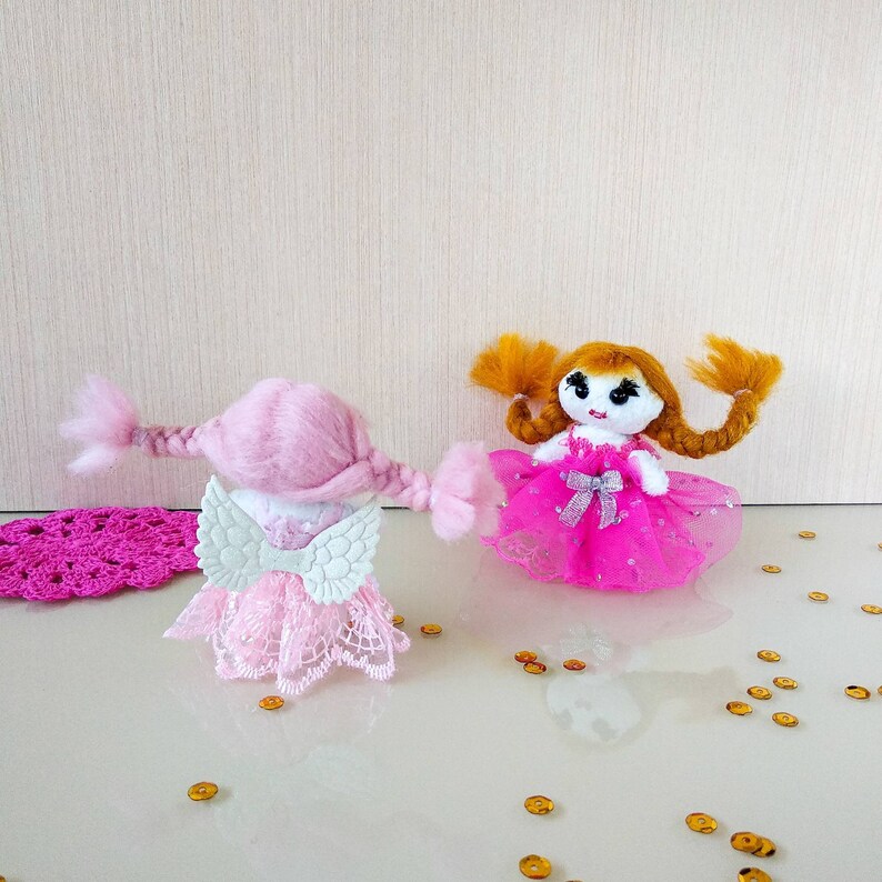 Op de afbeelding: Twee handgemaakte poppen. E&eacute;n pop is roze met witte vleugels en een kanten jurk. De andere pop heeft oranje haar en een roze jurk met een zilveren strik. Gouden confetti is verspreid rond de poppen.