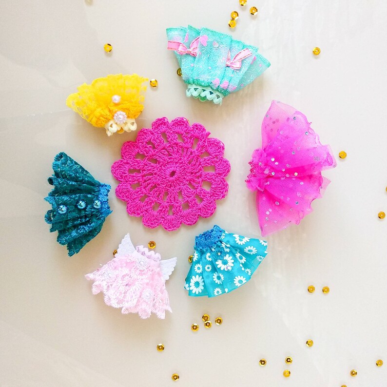 Op de afbeelding: Een verzameling kleurrijke poppenkleding en accessoires, waaronder minirokjes in teal, roze en blauw, een gele kanten hoofdband en een felroze gehaakte placemat. Gouden pailletten zijn rond de items verspreid.