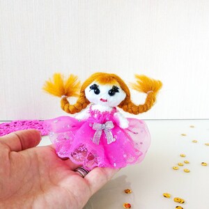 Op de afbeelding: Handgemaakte pop met oranje garen haar in twee vlechten. De pop draagt een roze jurk met een zilveren strik en een tule rok. Het gezicht van de pop heeft zwarte ogen en een rode mond. Ambachtelijk artikel.