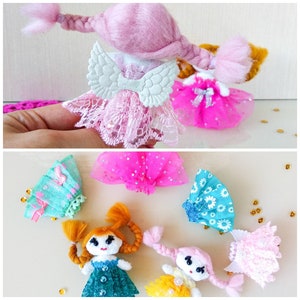 Op de afbeelding: Een verzameling handgemaakte poppenfiguren met garen haar en kleine jurkjes. De poppen hebben gevlochten haar in roze, geel en oranje. Sommige poppen hebben kanten jurkjes en zilveren vleugels. De jurkjes zijn in verschillende kleuren, waaronder roze, blauw en geel.