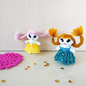 Op de afbeelding: Twee handgemaakte garendolls met lang gevlochten haar. De ene pop heeft roze haar en een gele jurk, de andere heeft oranje haar en een turquoise jurk. Beide poppen hebben zwarte ogen en zijn versierd met pailletten.