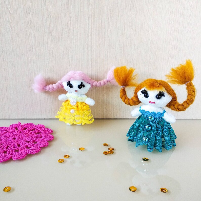 Op de afbeelding: Twee handgemaakte garendolls met lang gevlochten haar. De ene pop heeft roze haar en een gele jurk, de andere heeft oranje haar en een turquoise jurk. Beide poppen hebben zwarte ogen en zijn versierd met pailletten.