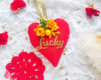 Könnte beinhalten: Rotes Filzherz-Anhänger mit goldenen Glitzerbuchstaben, auf denen "Lucky" steht. Das Herz ist mit gelben Stoffblumen und goldenen Perlen verziert.