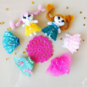 Op de afbeelding: Handgemaakte poppenfiguren met garen haar en stoffen jurken in geel, teal en roze. Een roze gehaakt kleedje en verschillende poppenrokken zijn ook aanwezig. Gouden pailletten zijn rond de poppen verspreid.